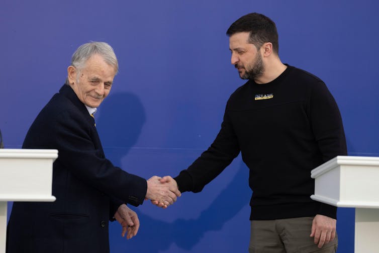 Ukrainian president Volodymyr Zelensky shakes hands with an older man, both stood at lecterns