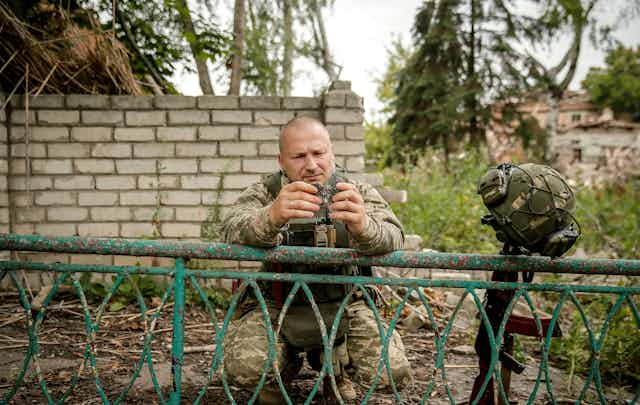 A Ukrainian soldier examines a piece of debris from a Russian shell while leaning on a fence. His helmet is hung nearby on top of his rifle.