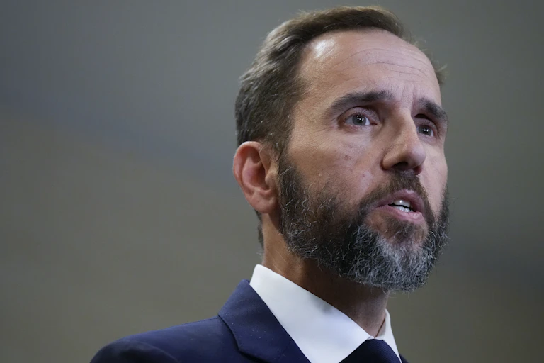 A white man with a beard and dressed in a business suit is talking to a group of people.