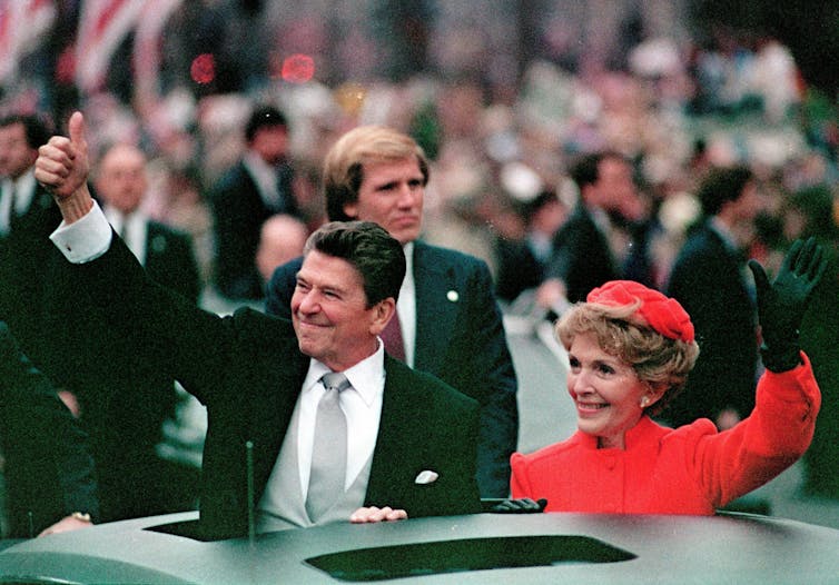 A dark-haired man gives a thumbs up while a woman dressed in red waves from a limousine.