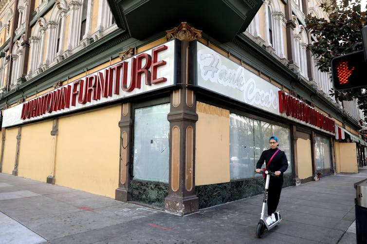 A young person rides a scooter past a shuttered store displaying the sign 
