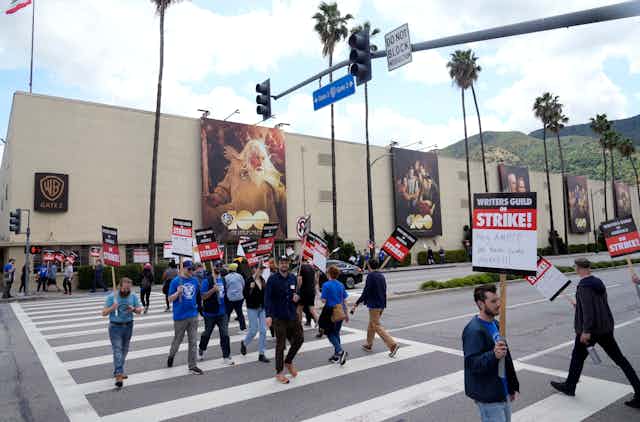 A group of people carrying signs walk in a crosswalk in front of a large building draped with large banners containing images of people