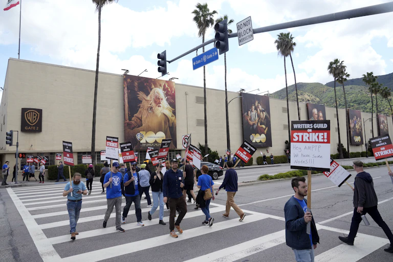 A group of people carrying signs walk in a crosswalk in front of a large building draped with large banners containing images of people