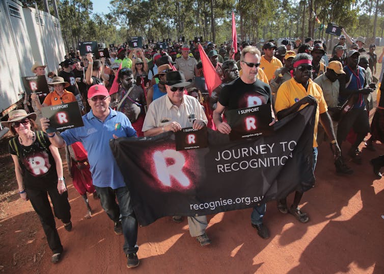 A group of people on a sunny day, walking with a flag supporting the Recognise campaign. They walk on red dirt.
