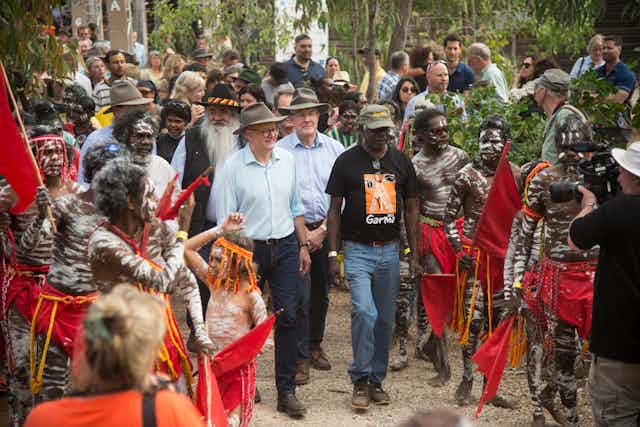 A white man, Prime Minister Albanese walks with Yolgnu man Djawa Yunupingu. They are surrounded by people, some are First Nations people in red lap-laps and painted in white ochre.