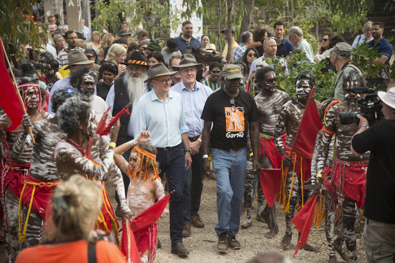 A white man, Prime Minister Albanese walks with Yolgnu man Djawa Yunupingu. They are surrounded by people, some are First Nations people in red lap-laps and painted in white ochre.