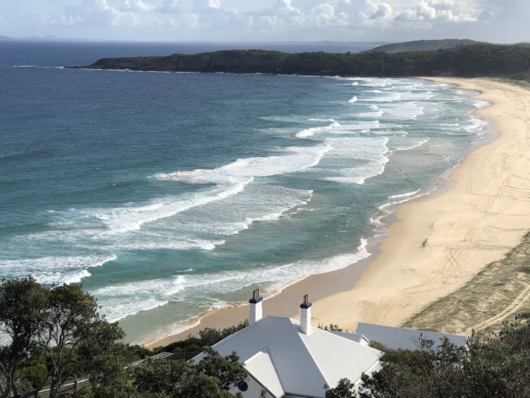 A view over a long, curved beach with rips visible at intervals among the waves