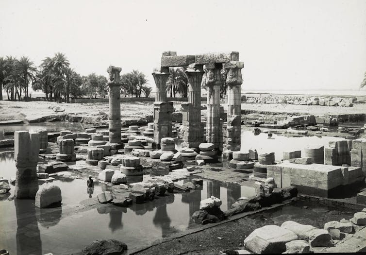 Imagen en blanco y negro. Se ven las ruinas de un templo egipcio con algunas columnas en pie. Todo el entorno está inundado y una persona asoma bañándose en el agua entre las ruinas.