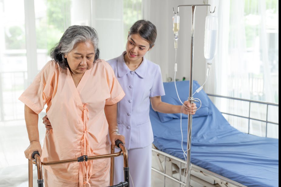 An older woman wearing a hospital gown uses a walker to exercise, while her nurse assists her.