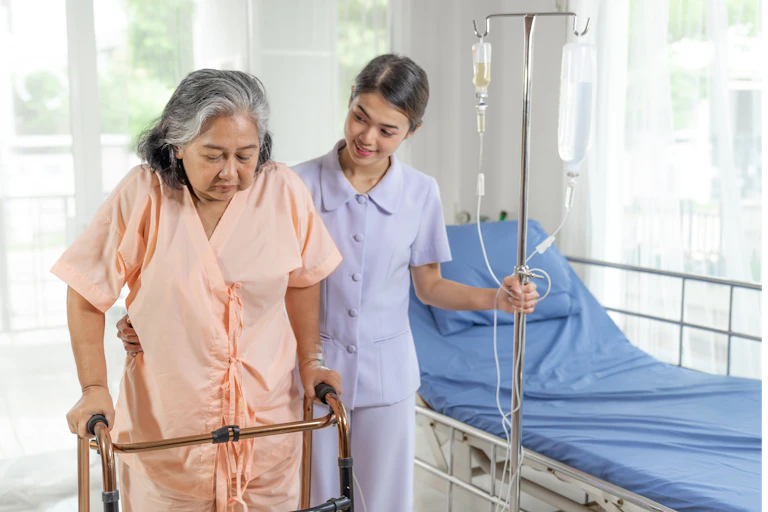 An older woman wearing a hospital gown uses a walker to exercise, while her nurse assists her.