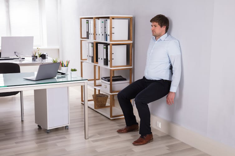 A man performs the wall sit exercise in his office.