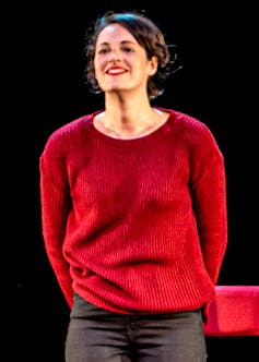 A young woman in a red jumper standing on a stage, spotlit.