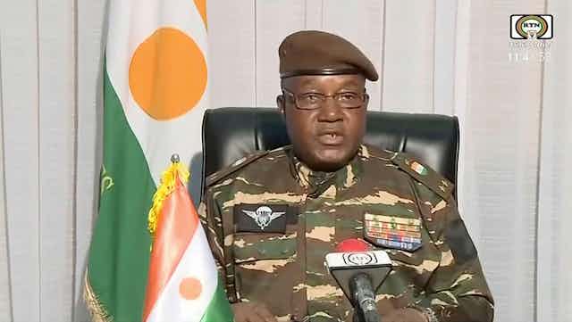 El general Abdourahmane Tchiani, líder golpista de Níger, con uniforme militar y flanqueado por la bandera del país.