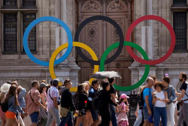 A crowd of people walking in front of the five Olympic rings displayed in front of a building with tall, ornate doors