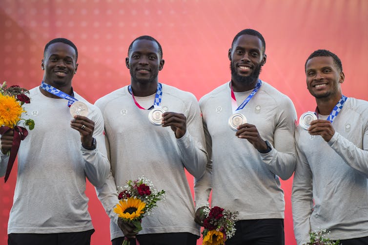 Four Black men in long-sleeve grey shirts smiling on a podium and holding up silver medals