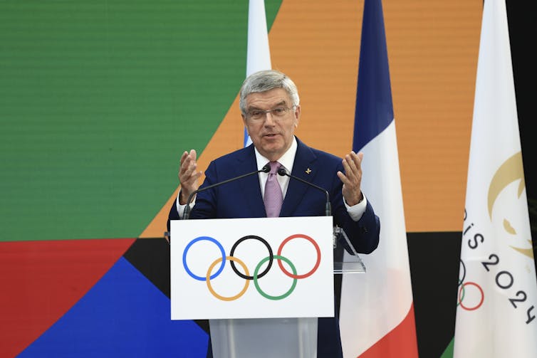 An older man with white hair and glasses gestures while speaking from behind a podium emblazoned with the Olympics logo