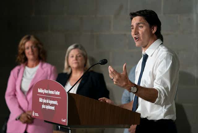 A middle-aged man in a white dress shirt and tie gestures while speaking from behind a podium that says 'Building More Homes Faster' on the front