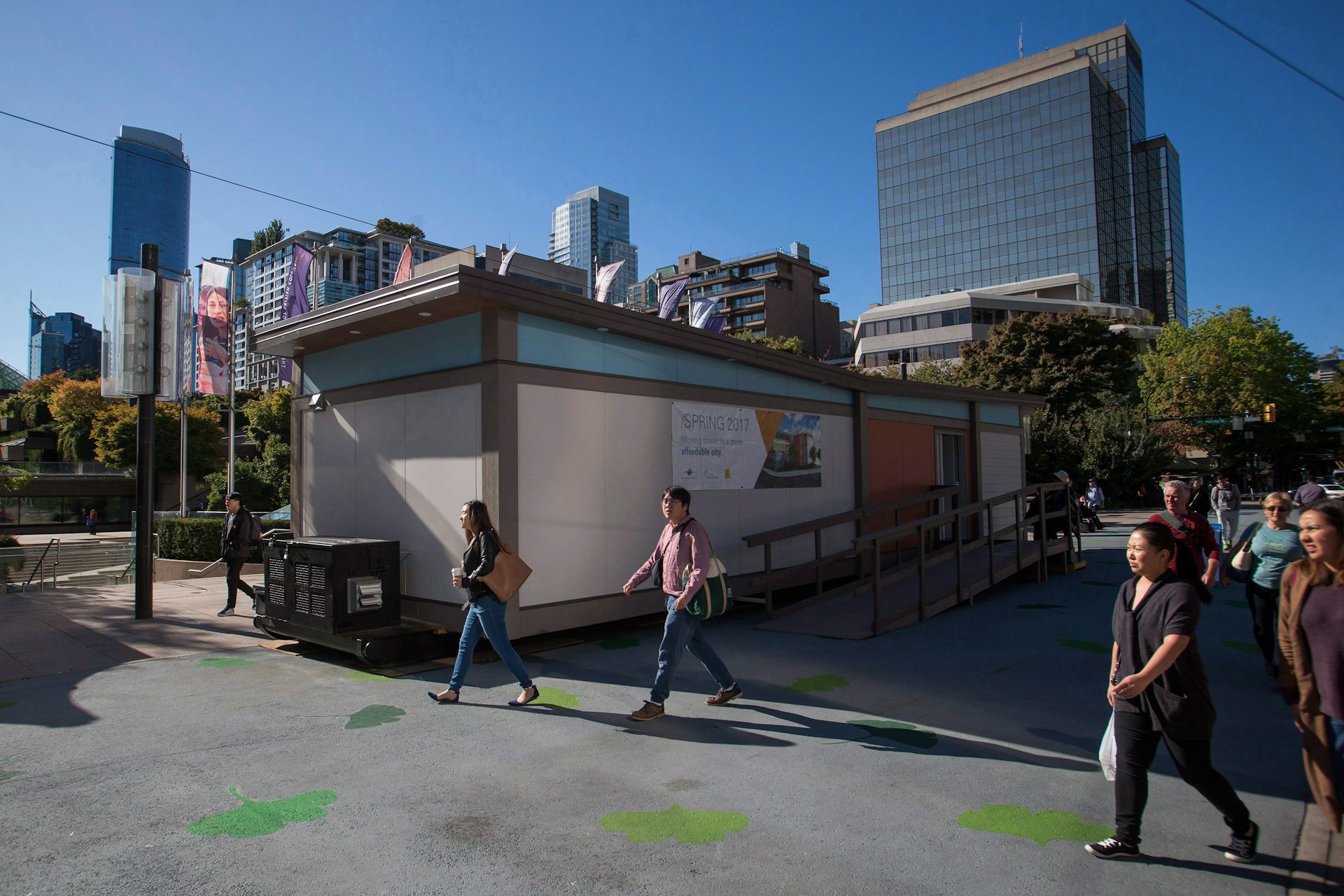 People walk past a small, rectangular residence. A city skyline is visible in the background.