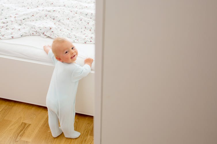 baby standing holding onto side of bed, looking back at camera