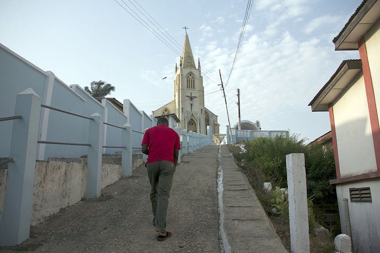 A man in a red shirt walks up a concrete walkway bordered by a fence, with a church at the top of the hill.