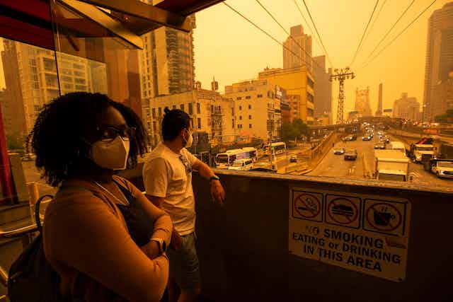 A woman wearing a face mask to protect against air pollution looks at the camera as tram moves into New York City from Roosevelt Island. The sky, with thick smoke particles reflecting sunlight,  is an orange color often used to suggest post-apocalyptic scenes in movies.