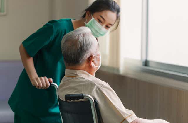 Aged care worker in a mask talks to an elderly patient in a wheelchair