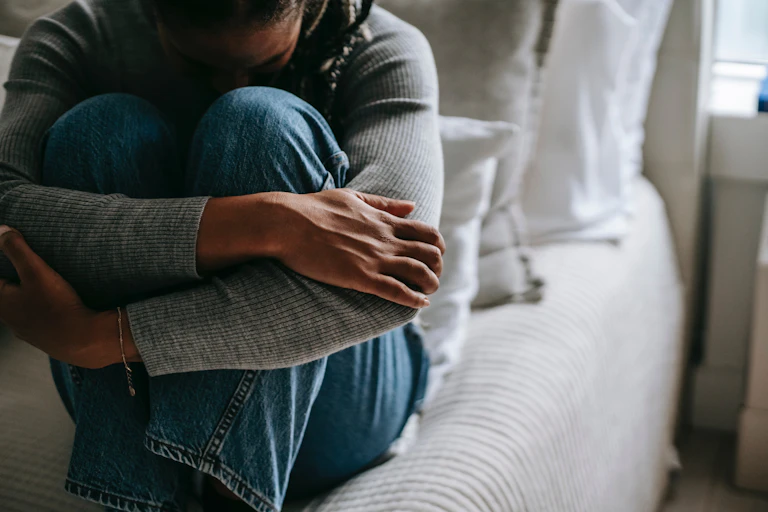 woman sits on bed, bows head, clasps knees