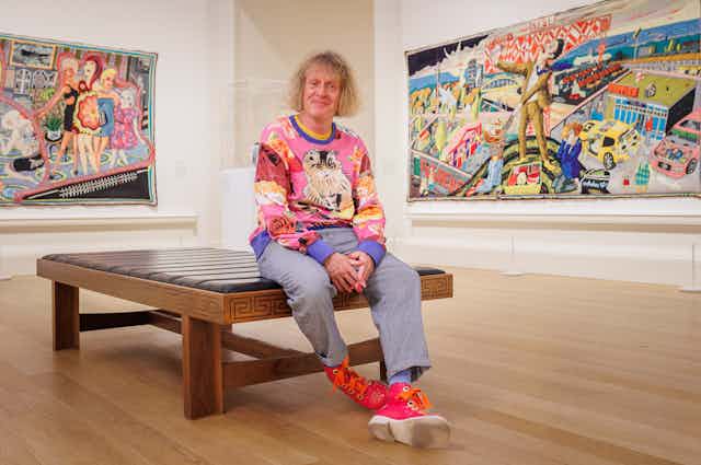 A blond man in a colourful jumper sitting on a bench in an art gallery.