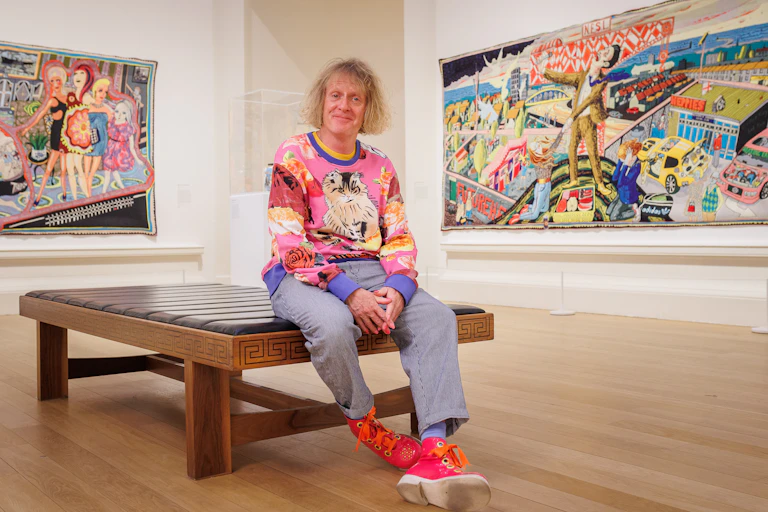 A blond man in a colourful jumper sitting on a bench in an art gallery.