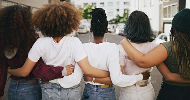 Five women of different races holding each other around the waists as they look towards a white building in the background with cars parked on the street in front of it