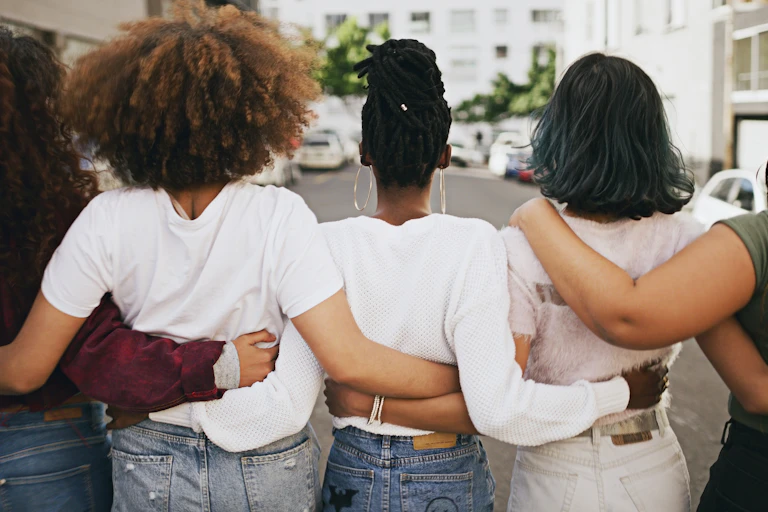 Five women of different races holding each other around the waists as they look towards a white building in the background with cars parked on the street in front of it
