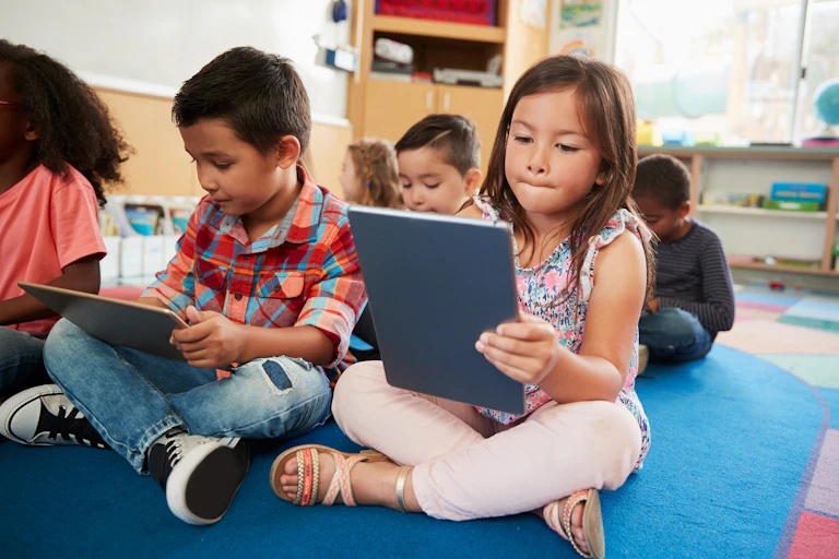 Dos niños sentados en el suelo de una clase consultan sus tabletas.