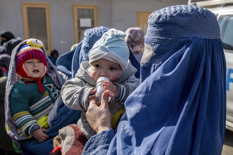 Uma mulher usando um niqab azul alimenta um bebê com uma mamadeira. Outro bebê está acenando ao fundo