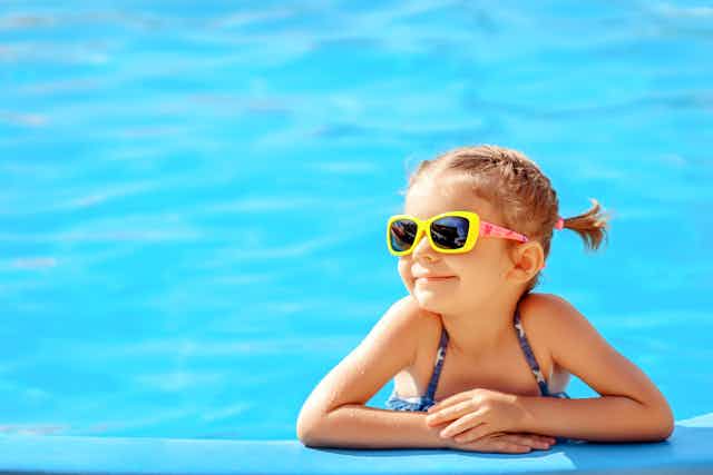 A small girl wearing sunglasses smiles from the border of a swimming pool.