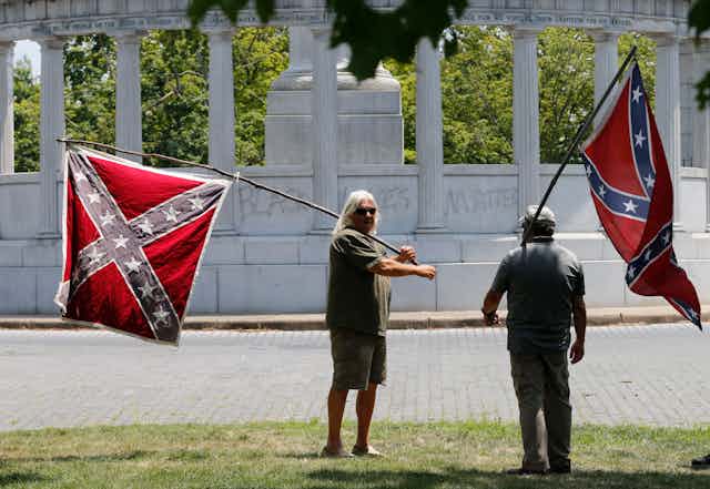 Two men standing outside in front of a large monument, holding Confederate flags.