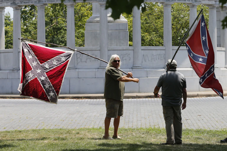 Two men standing outside in front of a large monument, holding Confederate flags.