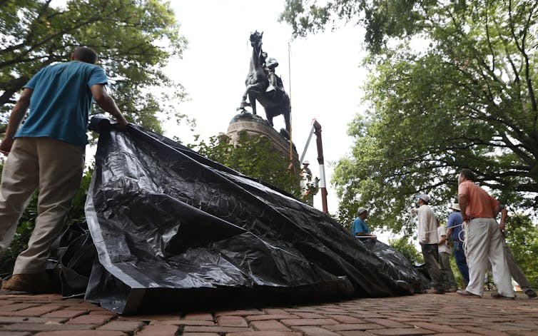 People hold a large tarpaulin beneath a statue of a man riding a horse.