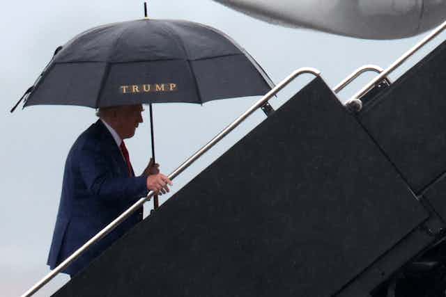 Donald Trump is seen walking up a flight of steps outside, holding a black umbrella with the word 'Trump' on it