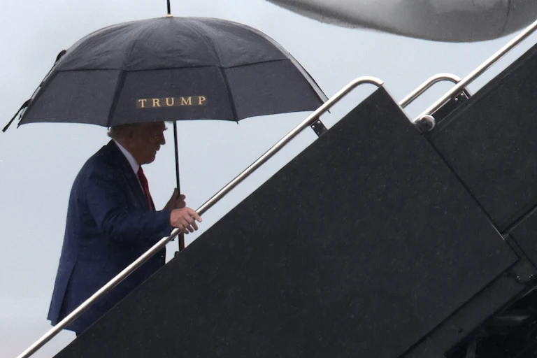 Donald Trump is seen walking up a flight of steps outside, holding a black umbrella with the word 'Trump' on it