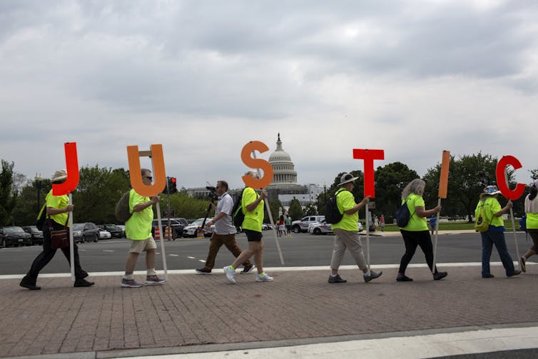 People wear neon green signs and hold up large letters spelling out the word 'justice' on a city street, in front of a building that looks similar to the U.S. Capitol.