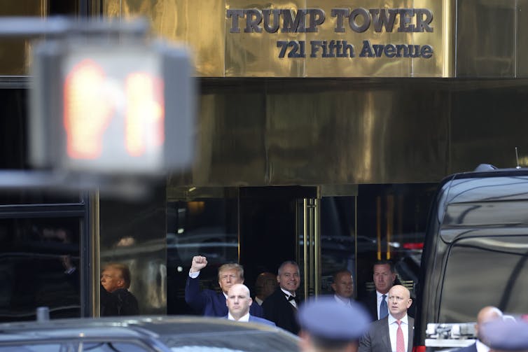 Donald Trump is seen pumping his fist in the air, benefath a Trump Tower sign in gold, and standing among other men in suits,