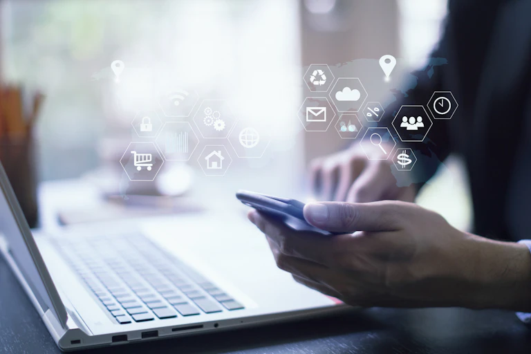 A hand holding a phone in front of a laptop. Many hexagonal icons float about the phone.