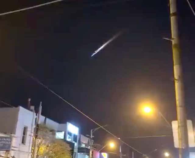 A grainy photograph showing a bright streak of light in the night sky above a city street.