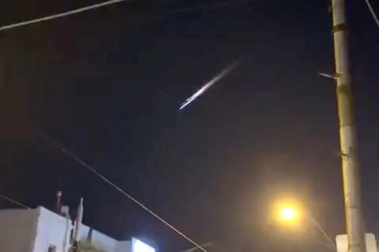 A grainy photograph showing a bright streak of light in the night sky above a city street.