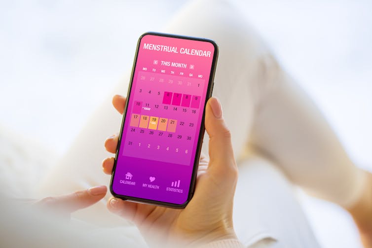 A hand holds a smartphone which is displaying a menstrual calendar app.