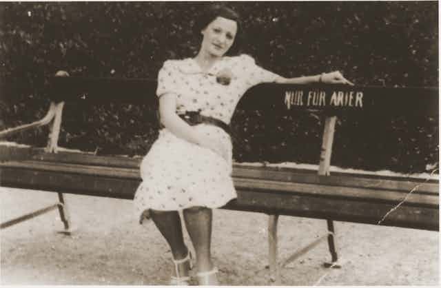 A dark-haired woman in a white print dress sitting on a bench outside that has a sign, in German, that says 'Only for Aryans.'