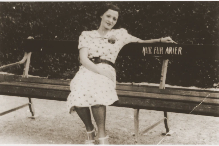 A dark-haired woman in a white print dress sitting on a bench outside that has a sign, in German, that says 'Only for Aryans.'
