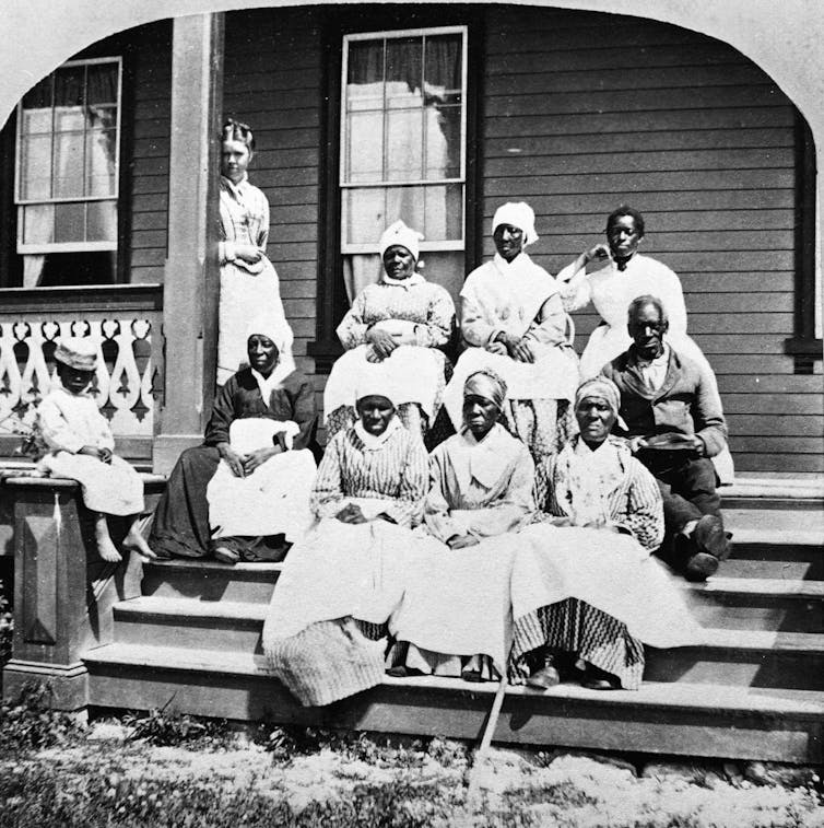 A group of enslaved African women and a man sit on the steps of a porch.