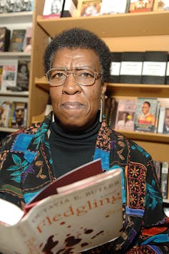 A woman with short hair, glasses and a brightly colored jacket stares into the camera, holding an open book.