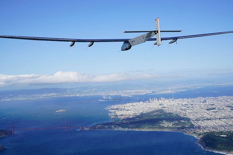 A futuristic airplane with a long wingspan flies over the bay and the city.